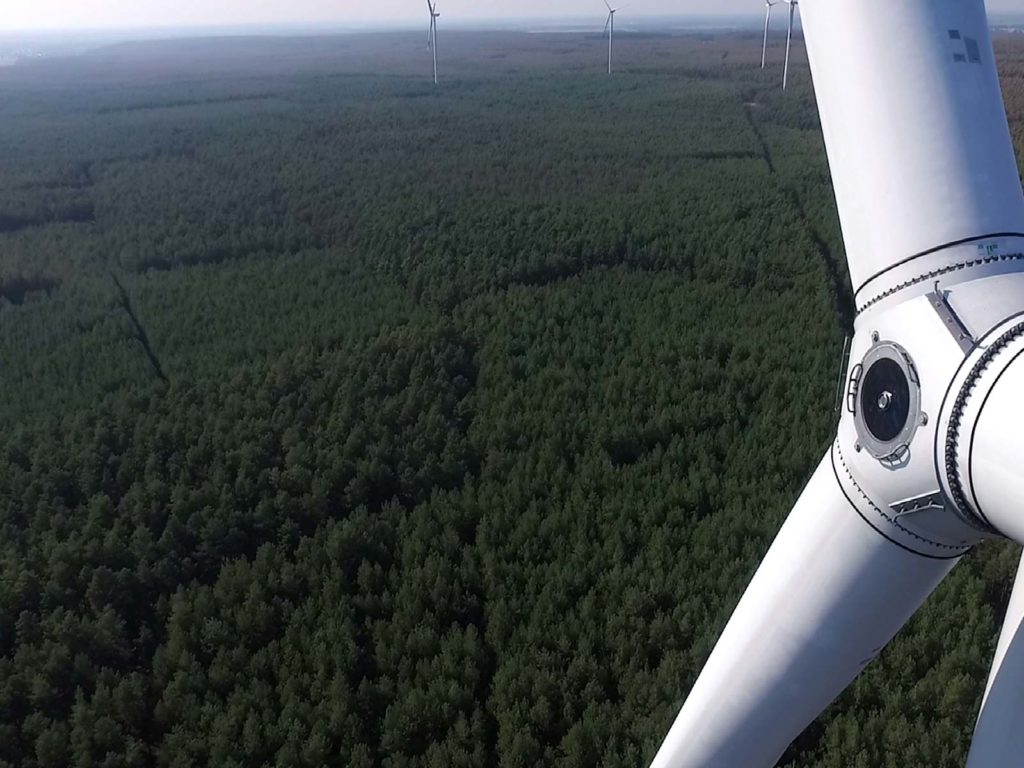 Luftbildaufnahme, auf der rechts ein Teil einer Windkraftanlage zu sehen ist und im Hintergrund Wald.